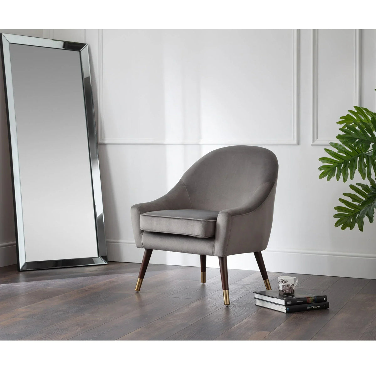 Grey Elliot armchair with wooden legs and gold tips in a modern room beside a tall mirror