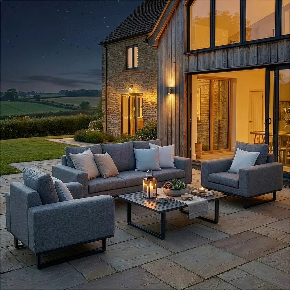 Grey outdoor sofa set with armchairs and cushions on stone patio, modern house in background