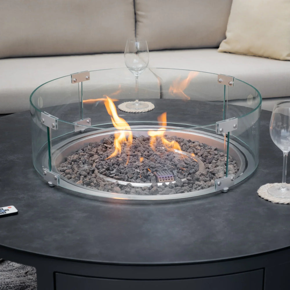 Round gas fire pit with glass wind guard on outdoor table, sofa and wine glasses nearby