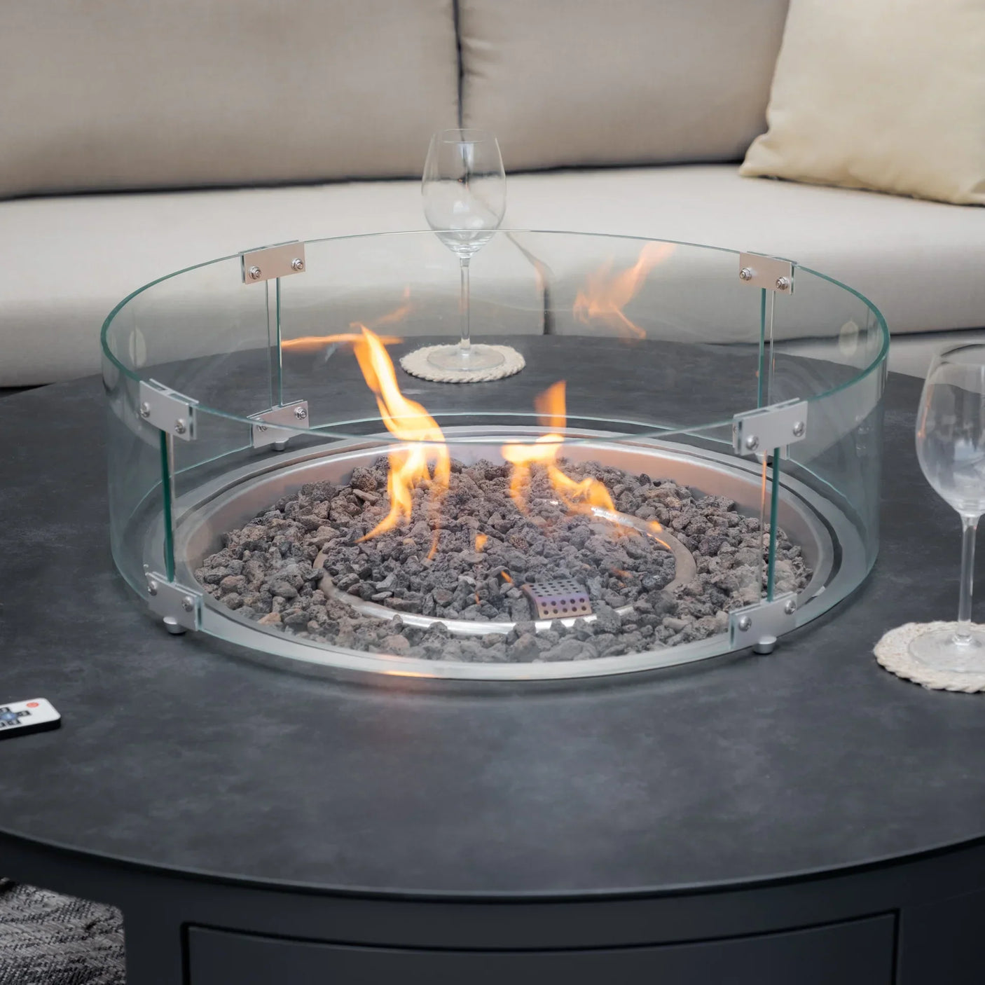 Round gas fire pit with glass wind guard on outdoor table, sofa and wine glasses nearby