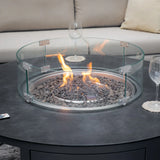 Round gas fire pit with glass wind guard on outdoor table, sofa and wine glasses nearby