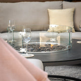 Round outdoor fire pit table with glass wind guard and lava rocks on patio seating area