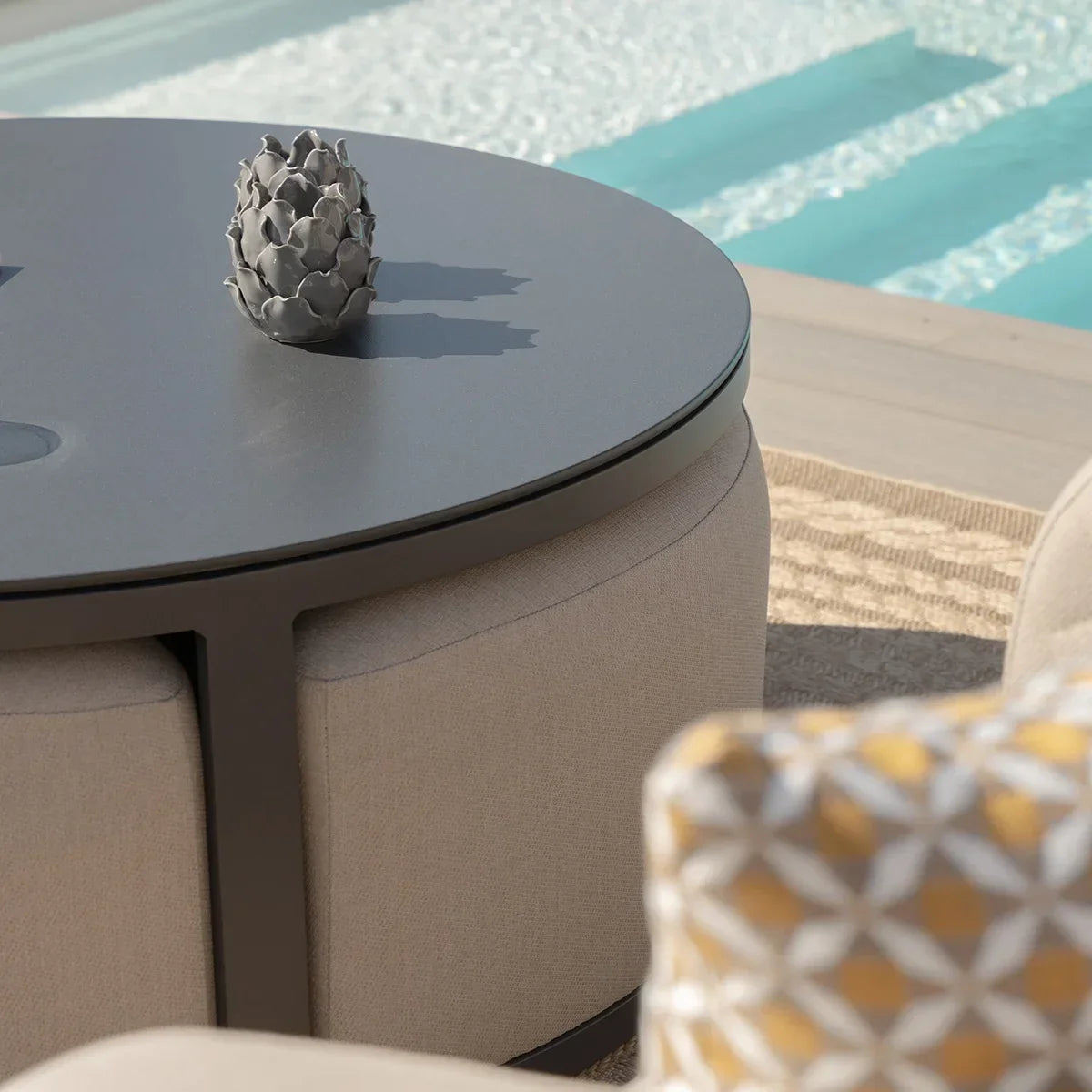 Round outdoor coffee table with dark top, beige ottomans and patterned cushion by pool