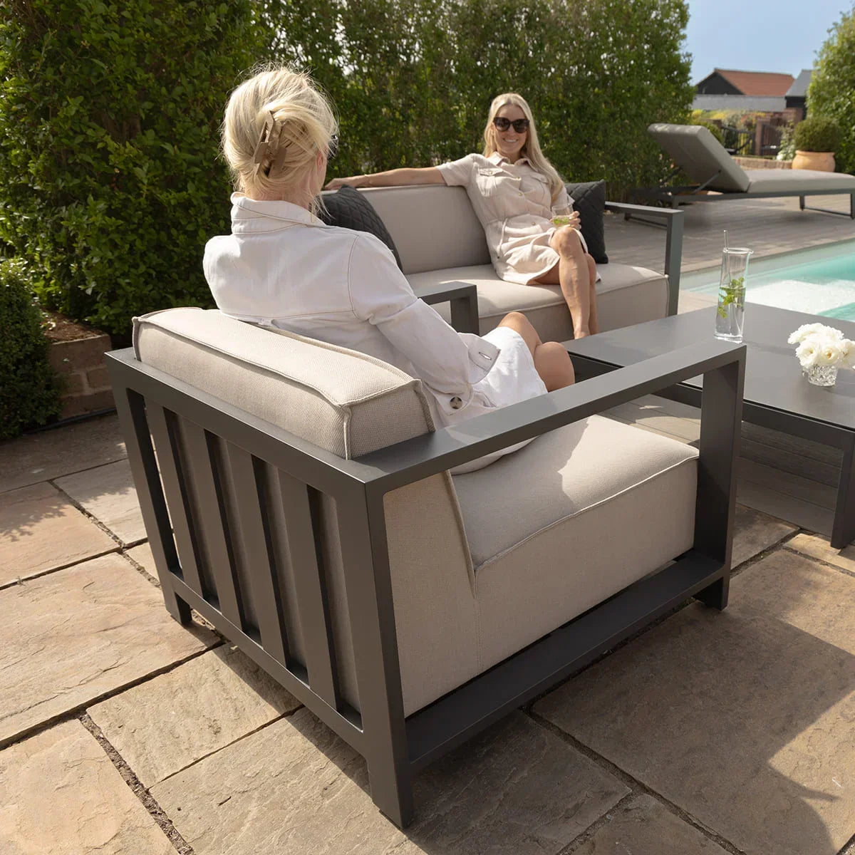 Grey metal outdoor armchair and sofa set beside pool on stone patio with two people seated