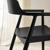 Side view detail of black wooden dining chair with angled legs and curved armrest near beige curtain.