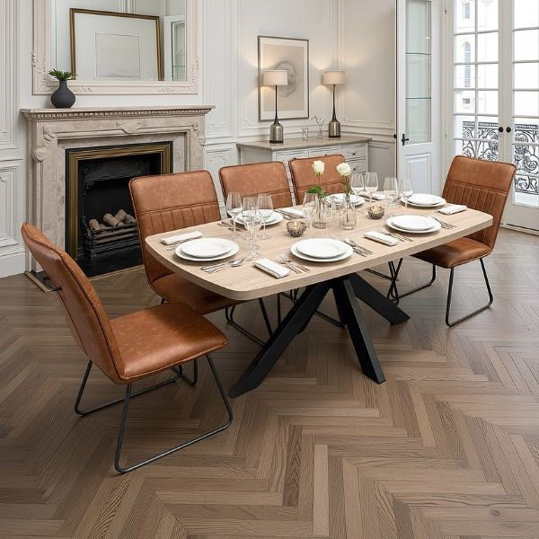 A modern dining room scene featuring a rectangular light wood dining table with black metal legs, surrounded by six brown leather upholstered chairs with black metal frames. The table is set with white plates, cutlery, and wine glasses, decorated with a small floral centerpiece.