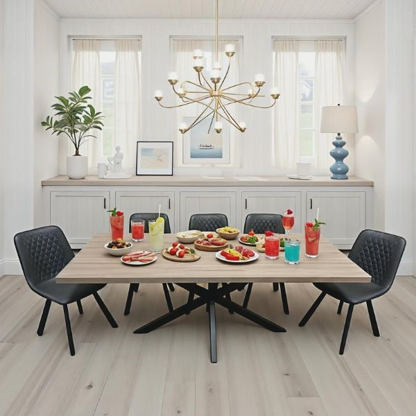 A modern dining room scene with a rectangular light wood table surrounded by six black quilted chairs. The table is set with colorful drinks, fruit, and small plates of food.
