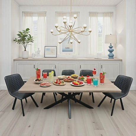 A modern dining room scene with a rectangular light wood table surrounded by six black quilted chairs. The table is set with colorful drinks, fruit, and small plates of food.