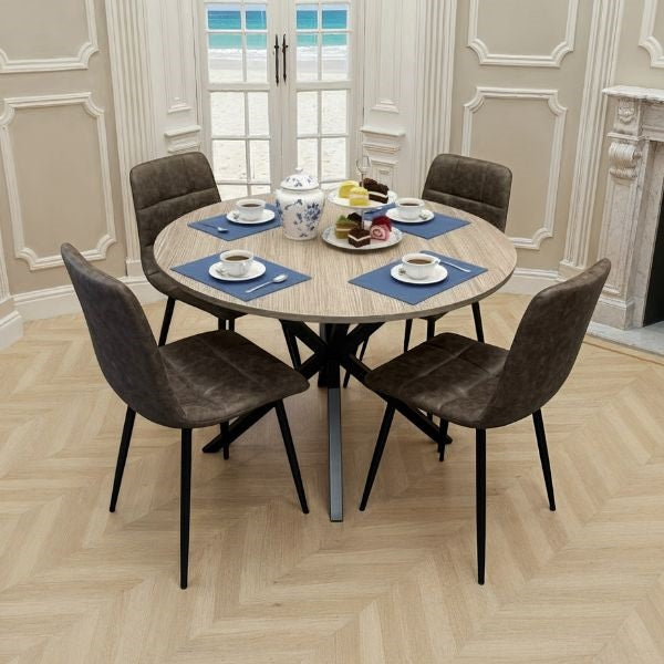 A round dining table with a light wood grain top and black metal pedestal base, surrounded by four dark brown upholstered chairs with black legs. The table is set with blue placemats, teacups, and a teapot in a bright dining room with beige walls and a herringbone wood floor.