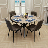 A round dining table with a light wood grain top and black metal pedestal base, surrounded by four dark brown upholstered chairs with black legs. The table is set with blue placemats, teacups, and a teapot in a bright dining room with beige walls and a herringbone wood floor.
