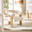 Cream boucle fabric accent armchair with rounded backrest, oak legs on a textured cream rug in bright room.