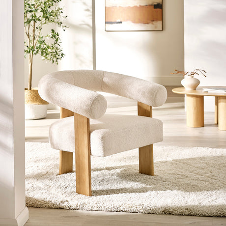 Cream boucle fabric accent armchair with rounded backrest, oak legs on a textured cream rug in bright room.