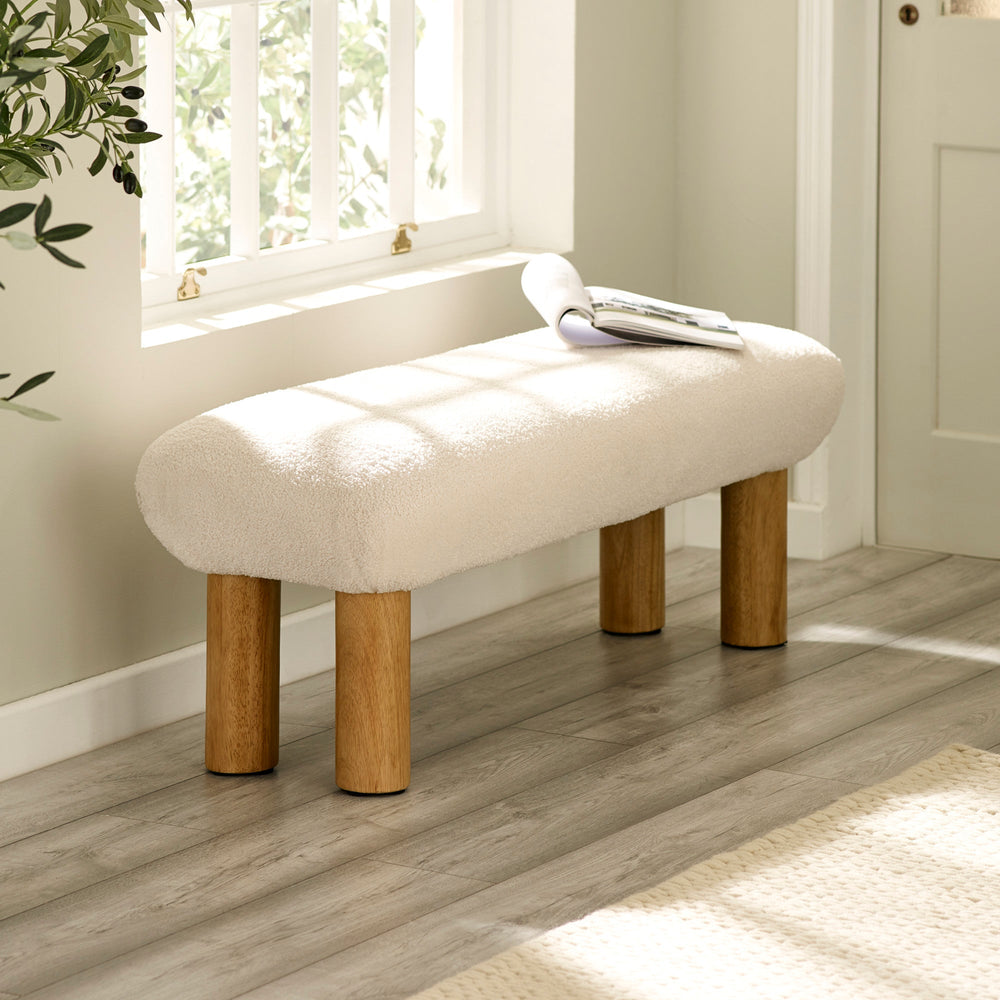 Cream upholstered low bench with cylindrical wooden legs beside a window with sunlight.