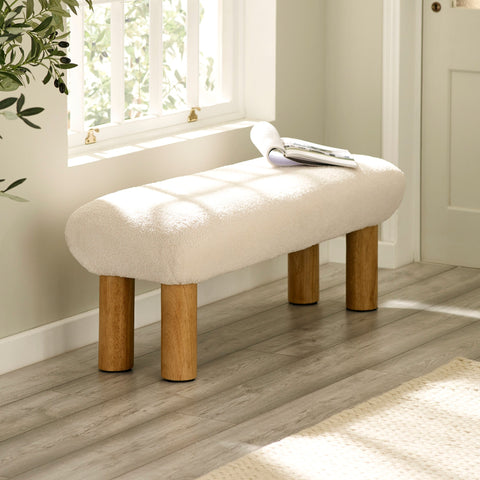 Cream upholstered low bench with cylindrical wooden legs beside a window with sunlight.