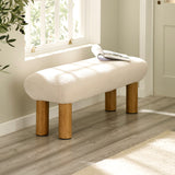 Cream upholstered low bench with cylindrical wooden legs beside a window with sunlight.