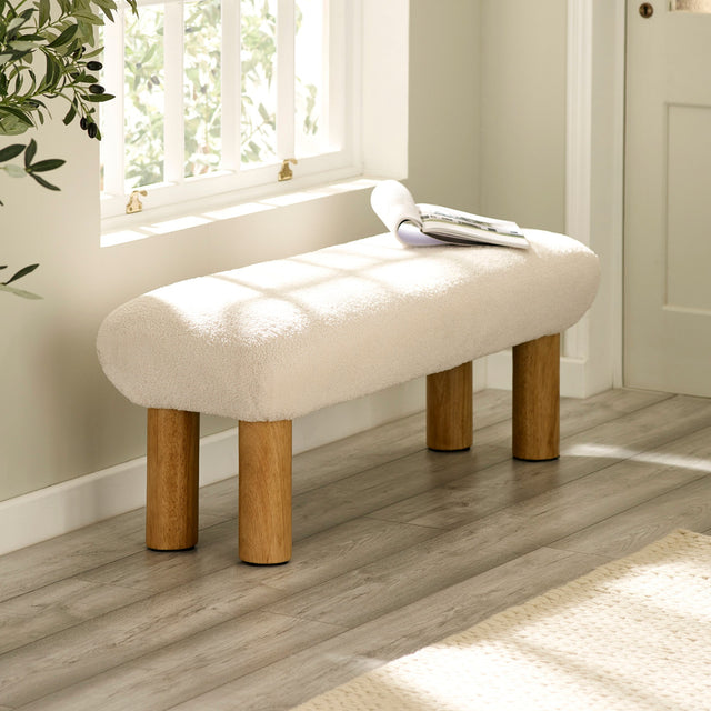 Cream upholstered low bench with cylindrical wooden legs beside a window with sunlight.