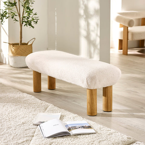 Front angle of cream low bench with four wooden legs on light hardwood floor near cozy rug and magazines.