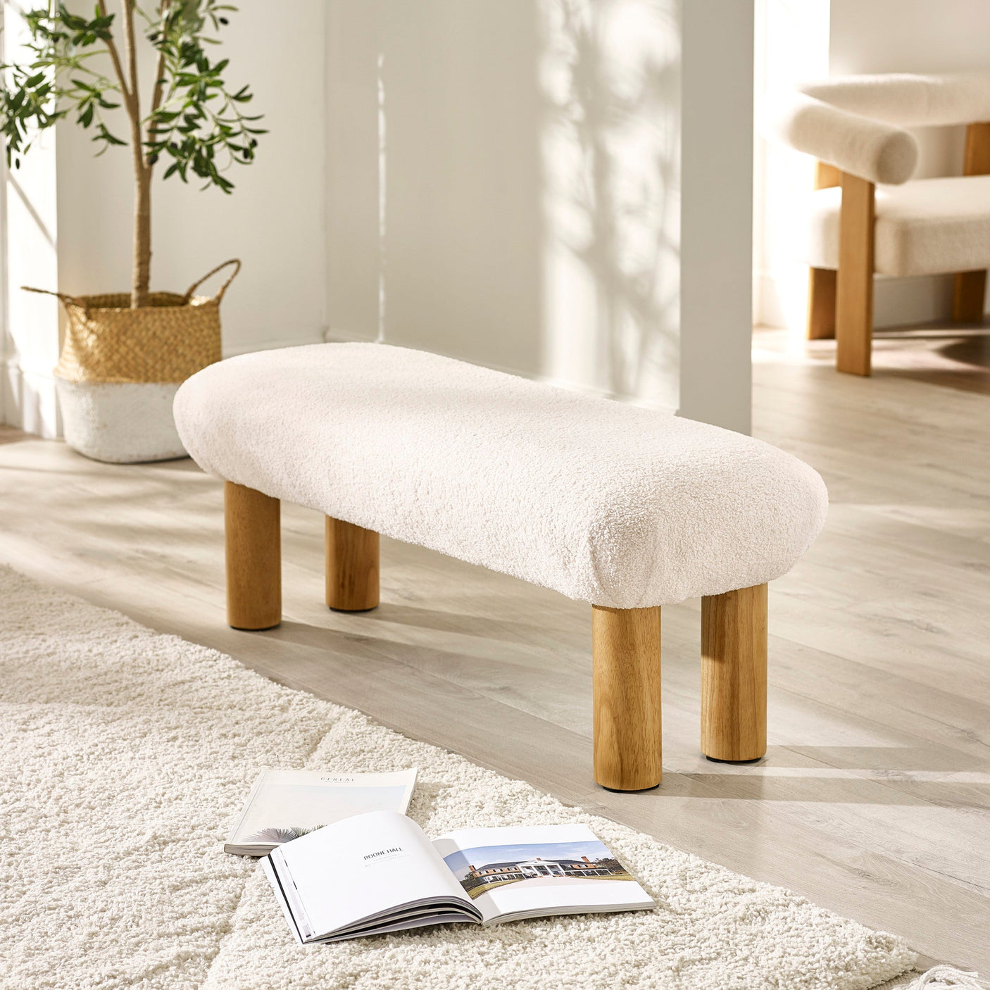 Front angle of cream low bench with four wooden legs on light hardwood floor near cozy rug and magazines.