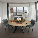 A round wooden dining table with black metal legs is set in a modern open-plan apartment. The table is styled with a Christmas wreath, white pillar candles, string lights, and holiday crackers, surrounded by four dark gray tufted chairs.