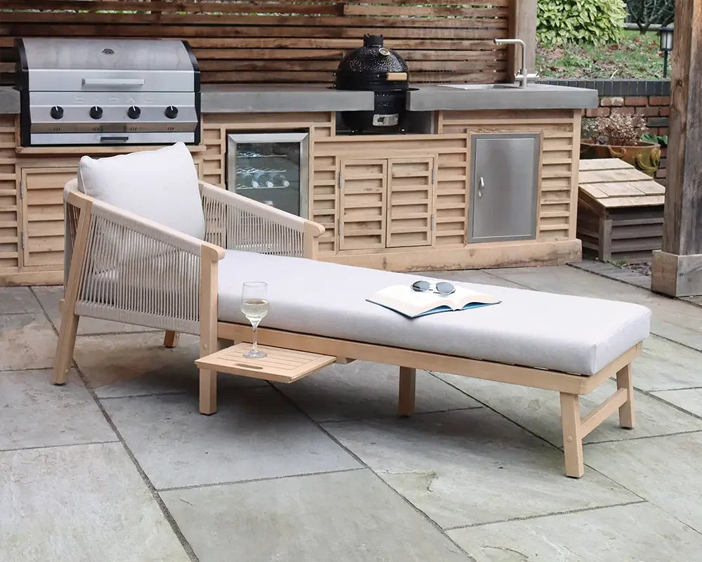 Outdoor wooden sun lounger with light cushion set on patio near barbecue and kitchenette