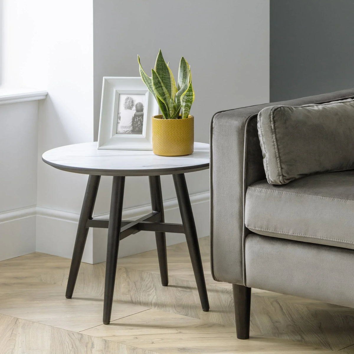 Round marble top lamp table with black legs beside grey sofa, plant and photo frame on top