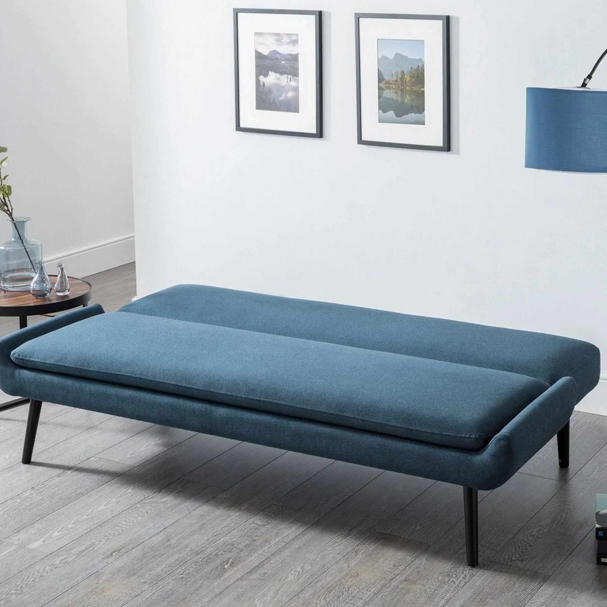 Blue sofabed with black legs in modern room with wood floor and two framed wall prints