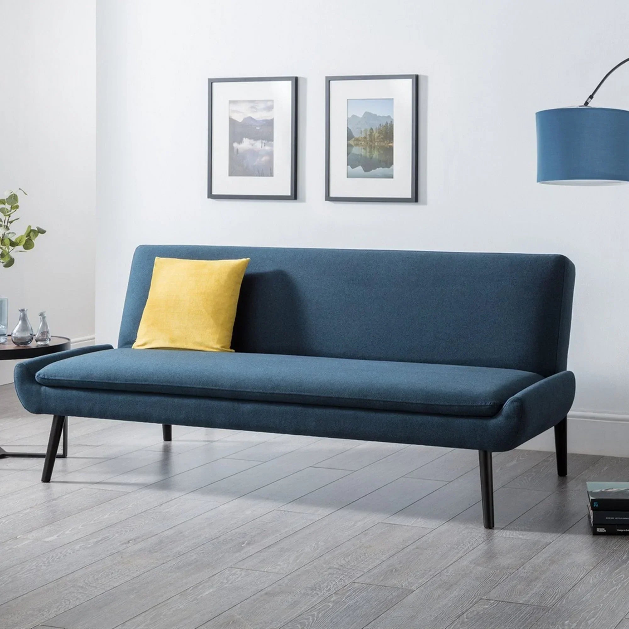Blue sofabed with angled legs in modern living room, yellow cushion, wall art in background