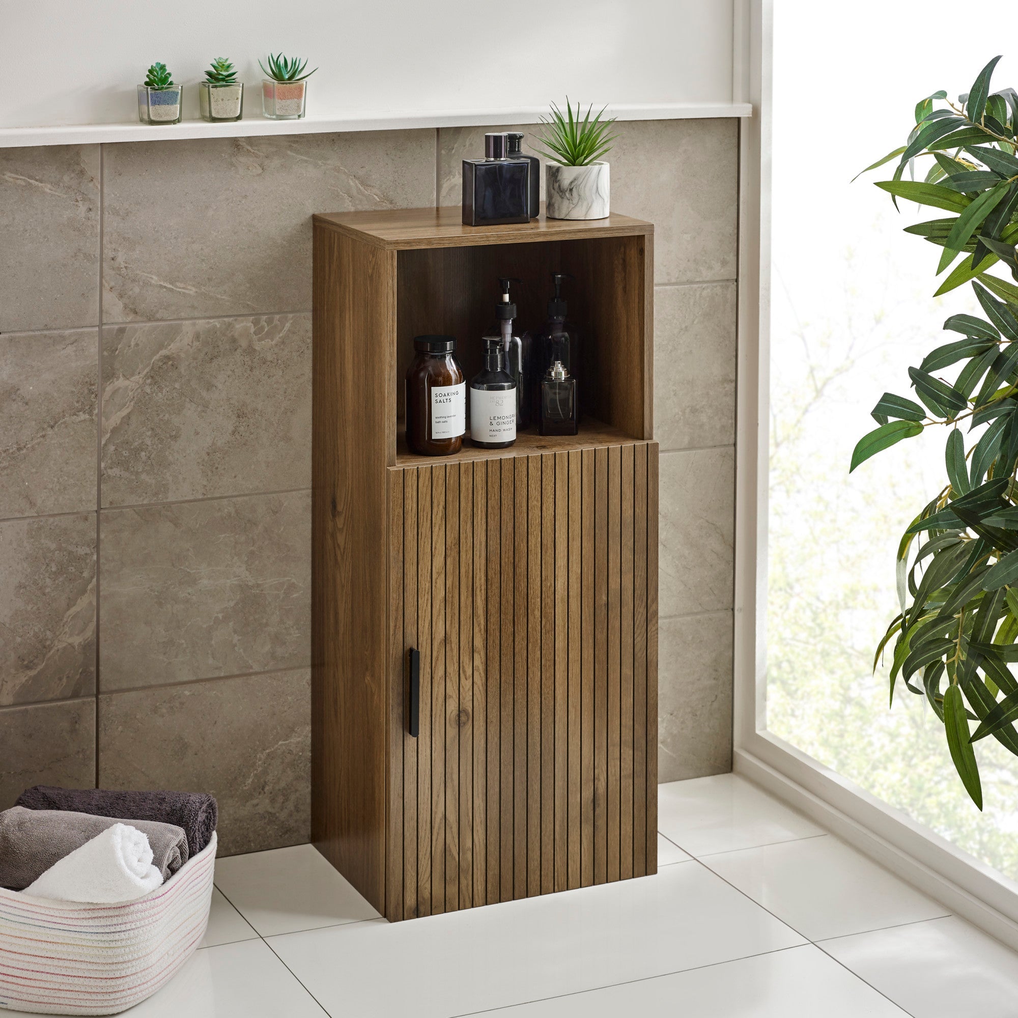 Tall brown wooden bathroom storage cabinet with an open top shelf holding bottles, side view in tiled bathroom.