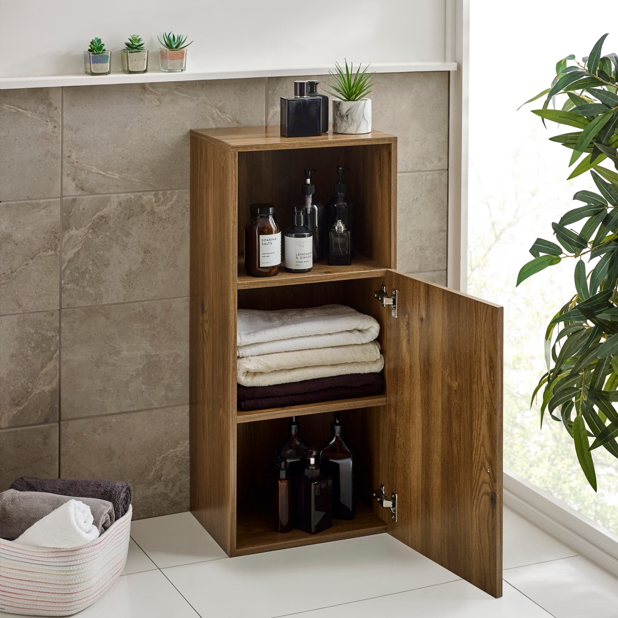 Brown wooden bathroom cabinet with one door open showing folded towels inside, next to window and plant.