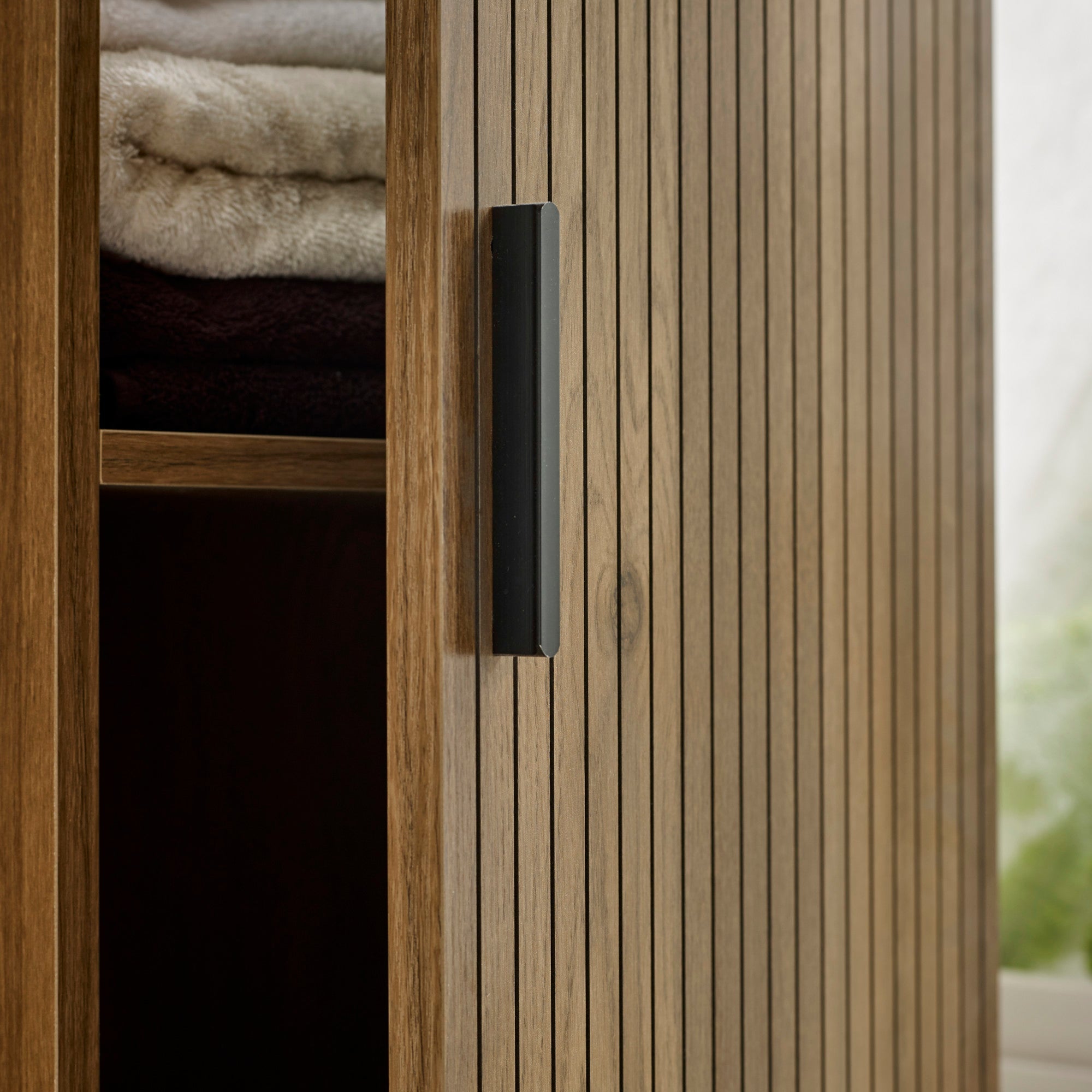 Detail of cabinet door with vertical grooves and black handle, partially opened with towels inside.
