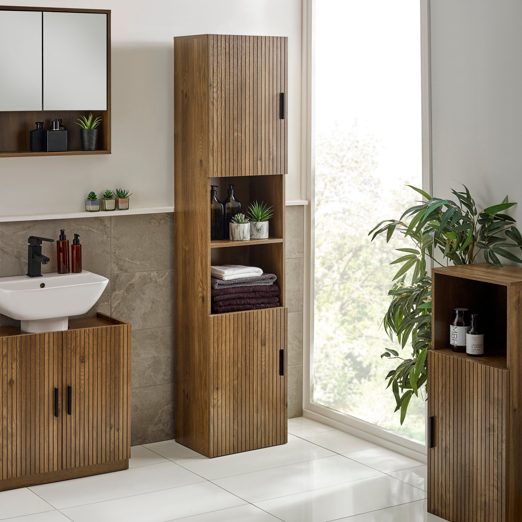 Tall bathroom cabinet with slatted wood doors, walnut finish, open middle shelves with plants and towels, near sink