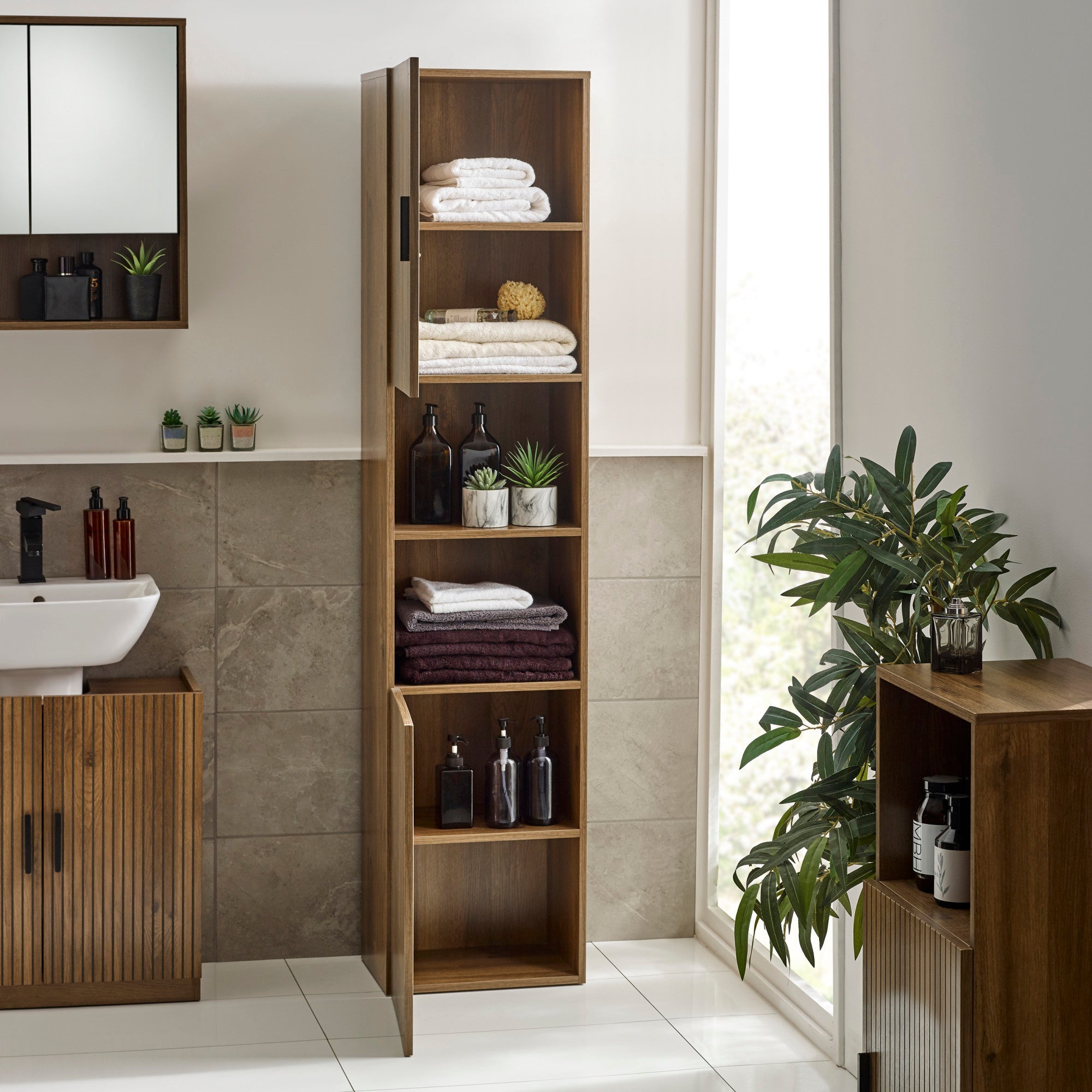 Bathroom cabinet with open doors showing stacked towels and bottles on wood shelves, warm walnut finish, tiled wall