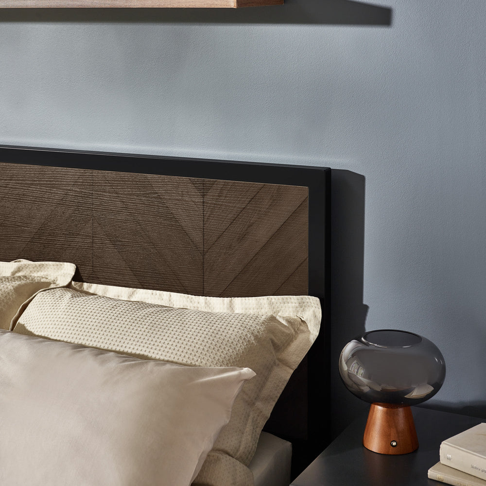 Angled view of dark wood herringbone headboard with cream pillows and bedside lamp on dark gray table
