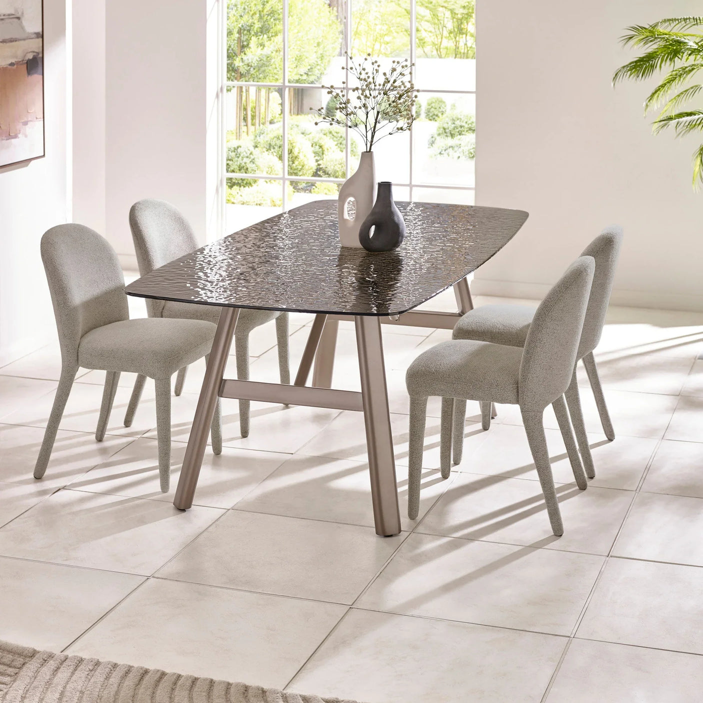 Glass dining table with taupe legs and four grey upholstered dining chairs in bright room