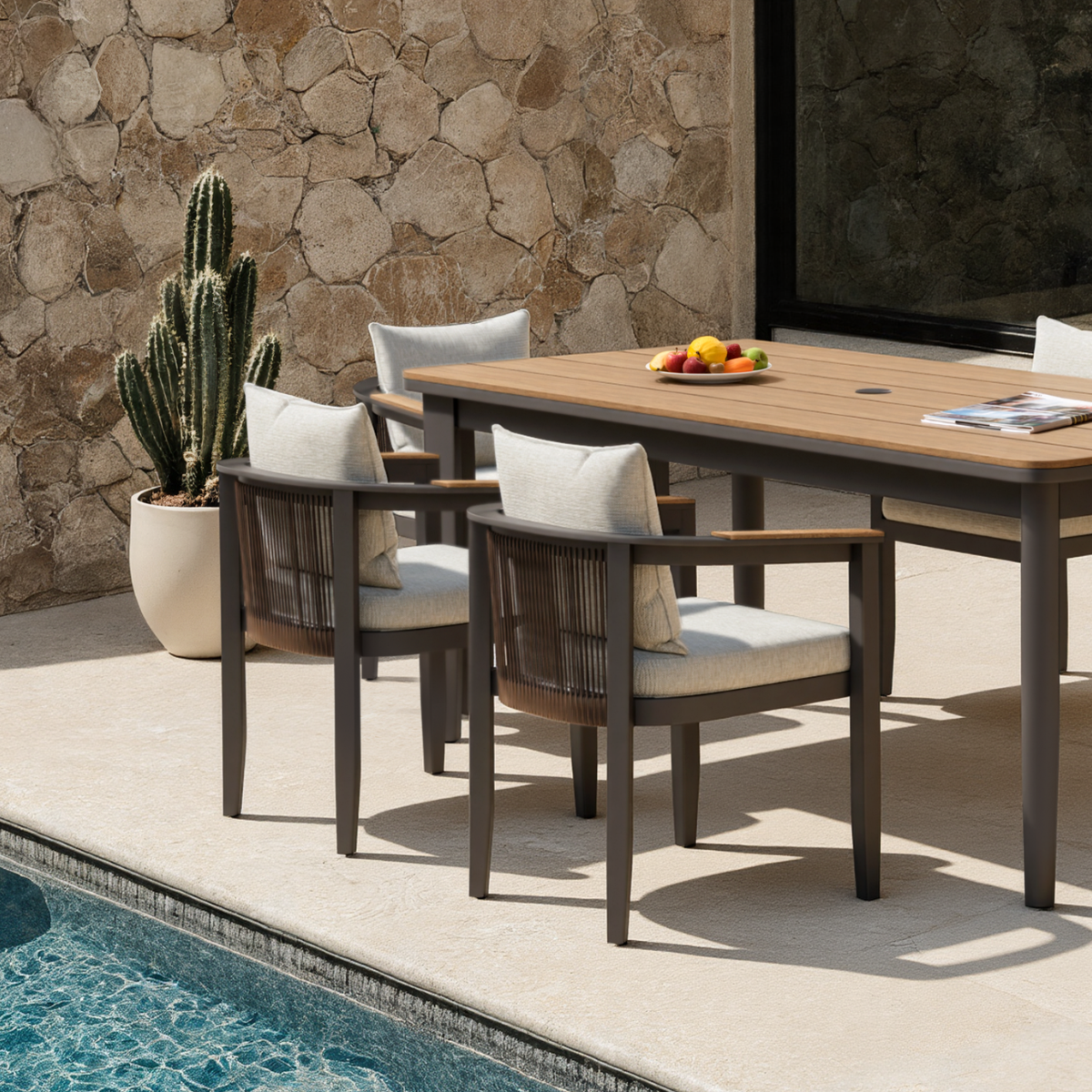 Light wood tabletop with dark grey frame chairs and beige cushions by a poolside.