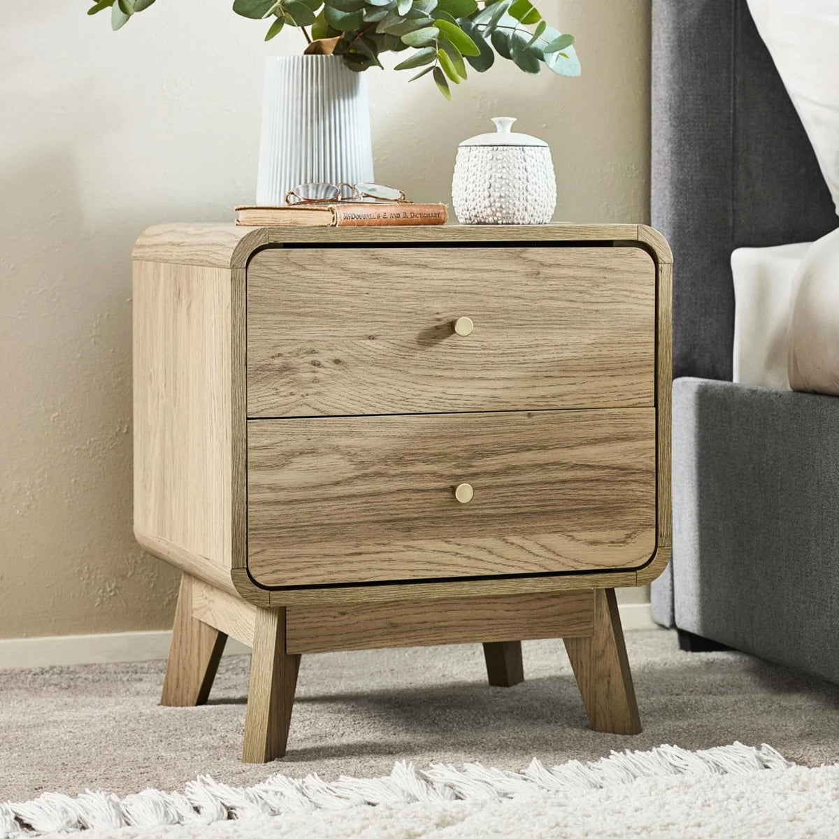 Oak bedside table with two drawers beside grey bed, styled with vase and decor items