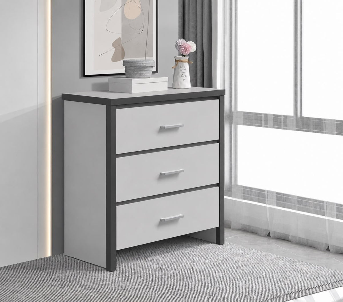 A modern three-drawer chest of drawers with a white finish and dark gray frame, styled in a bright, contemporary room next to a window.