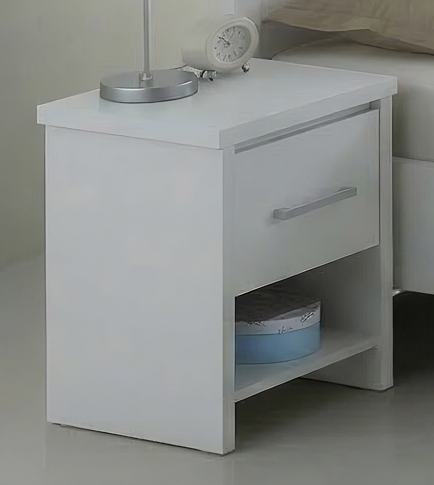 A white wooden nightstand with a single drawer and an open lower shelf, shown in a bedroom setting next to a bed. Decorative items on top include a silver desk lamp and a small white alarm clock.