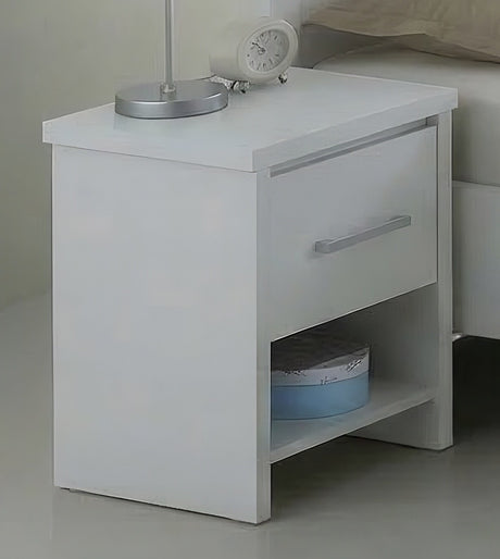 A white wooden nightstand with a single drawer and an open lower shelf, shown in a bedroom setting next to a bed. Decorative items on top include a silver desk lamp and a small white alarm clock.