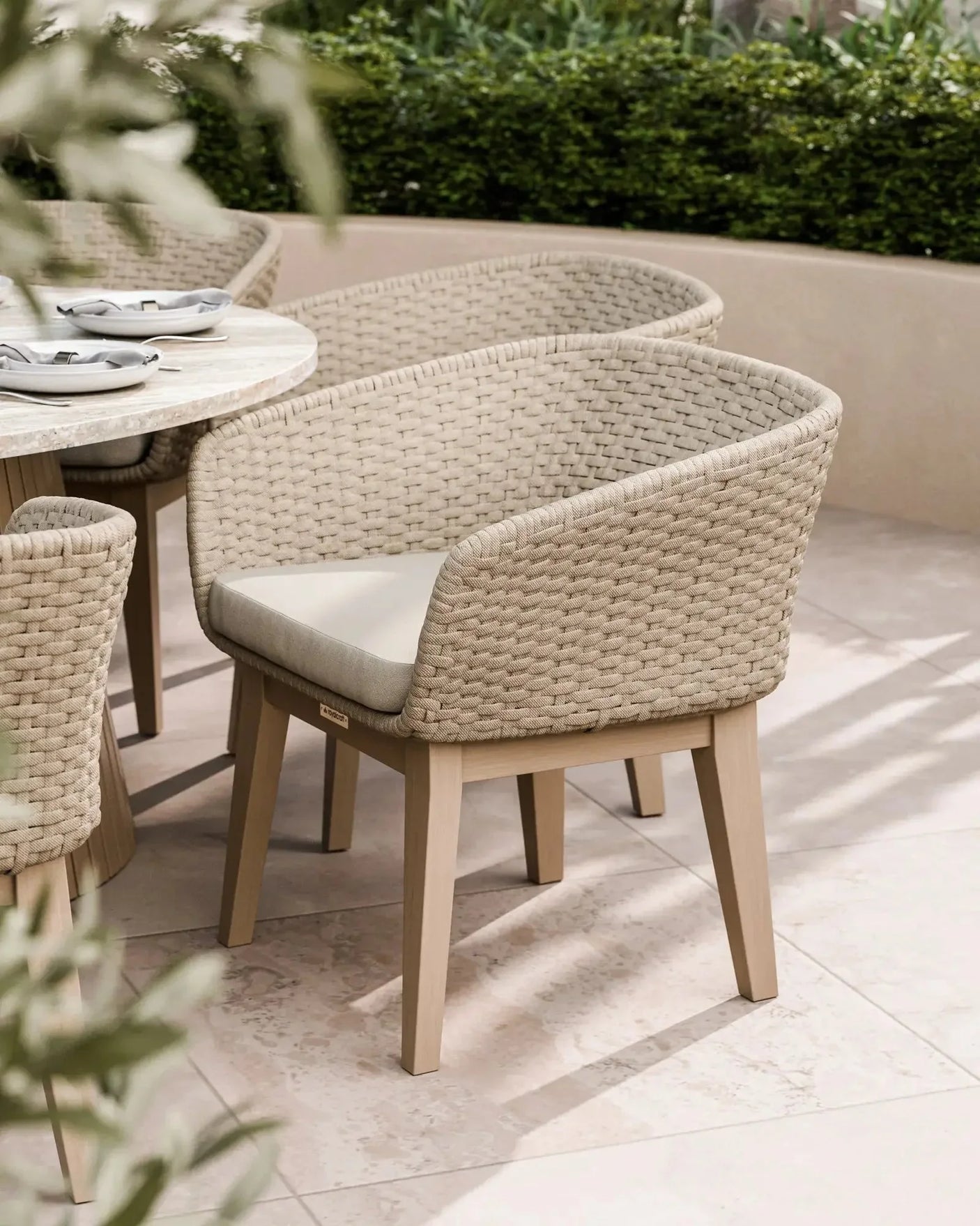 Rattan outdoor dining chair with light wood legs and cream cushion on tiled patio