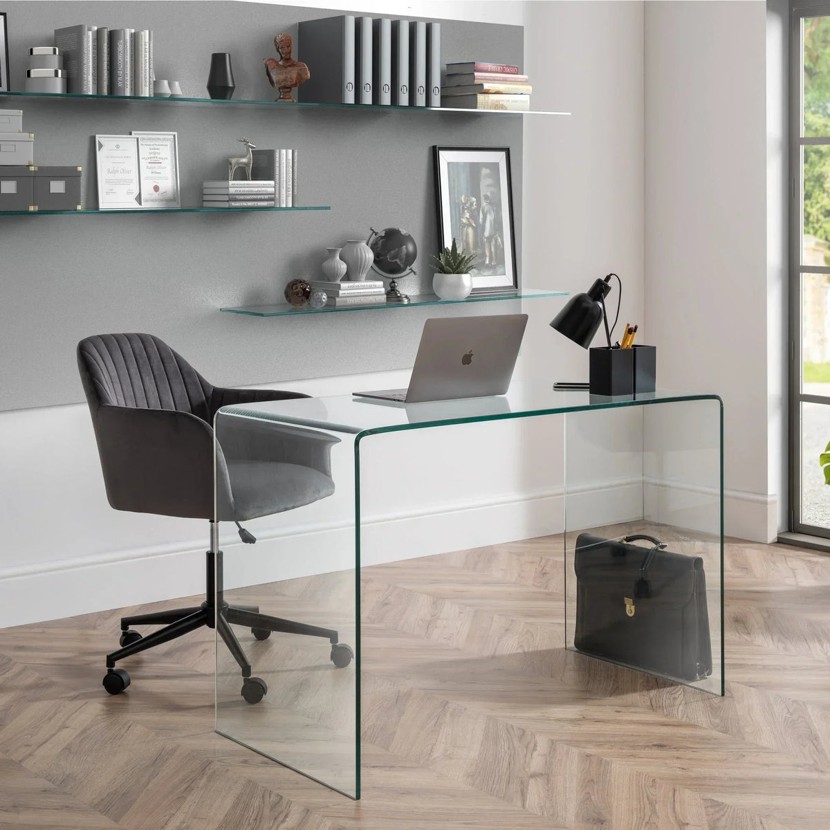 Grey office chair with armrests and wheels next to glass desk in modern workspace