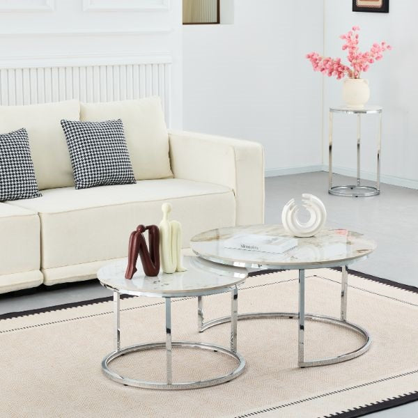 Two round nesting coffee tables with white marble-patterned tops and chrome metal bases, styled in a bright living room next to a cream sofa.