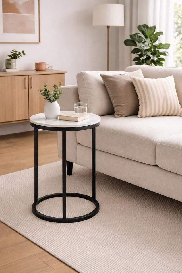 A round side table with a white top and black metal frame, styled in a modern living room next to a beige sofa.