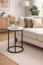A round side table with a white top and black metal frame, styled in a modern living room next to a beige sofa.