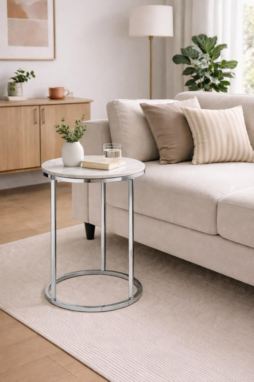 A round side table with a white top and a chrome metal frame, styled in a modern living room next to a beige sofa.
