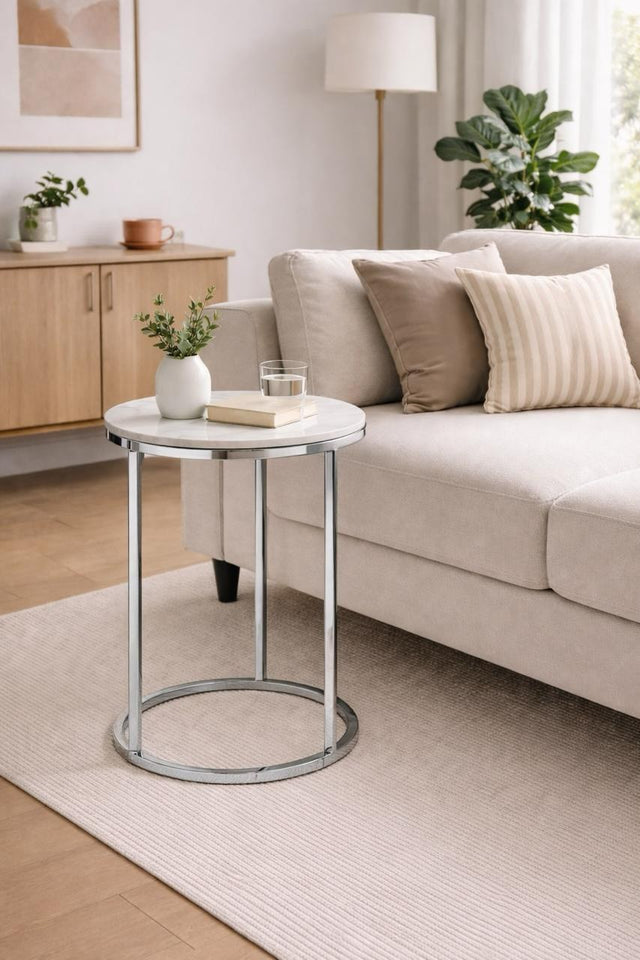 A round side table with a white top and a chrome metal frame, styled in a modern living room next to a beige sofa.