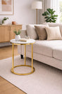 A round side table with a white marble top and a gold metal frame, styled in a modern living room next to a beige sofa.