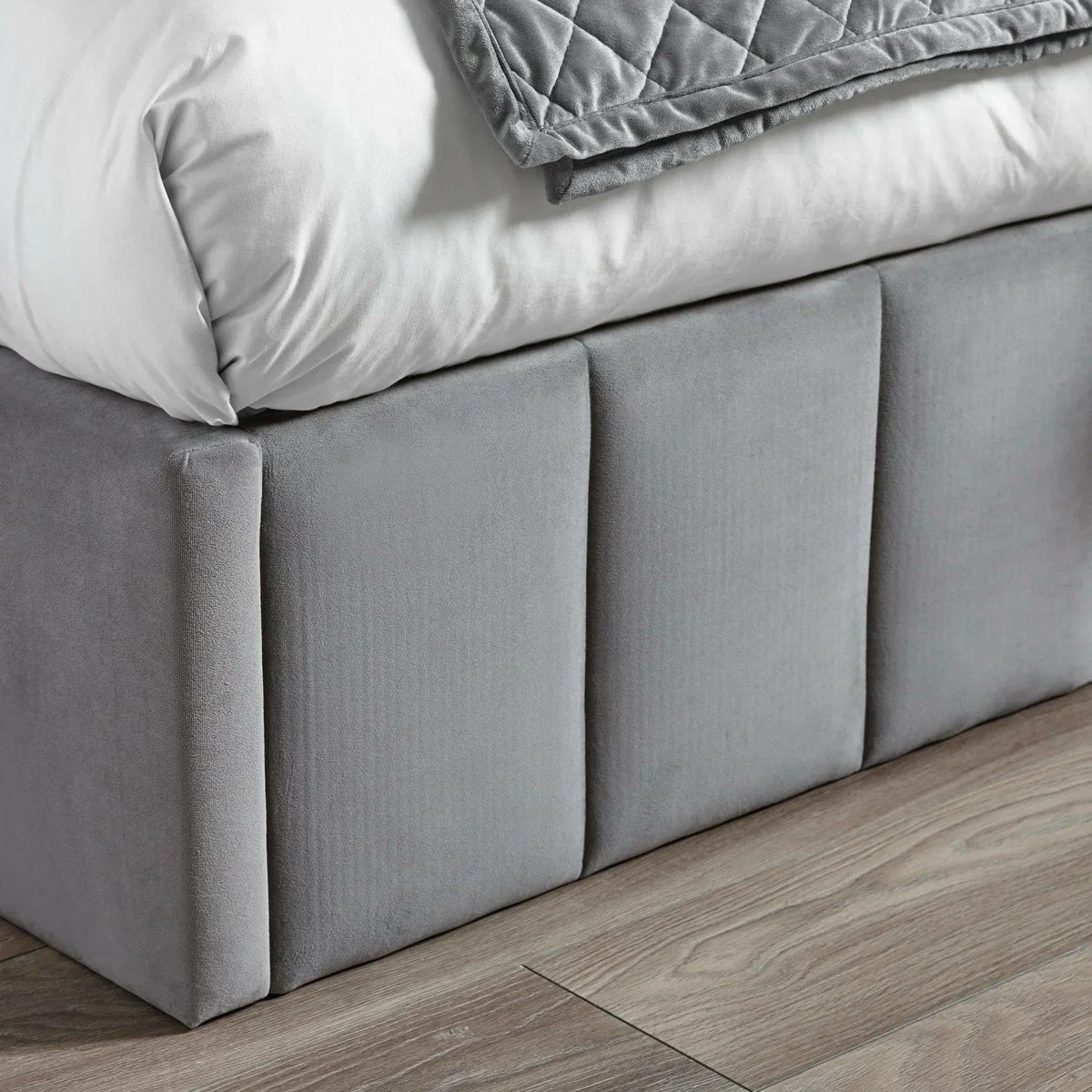 Grey velvet bed base with panel detail on wooden floor, partial bedding visible