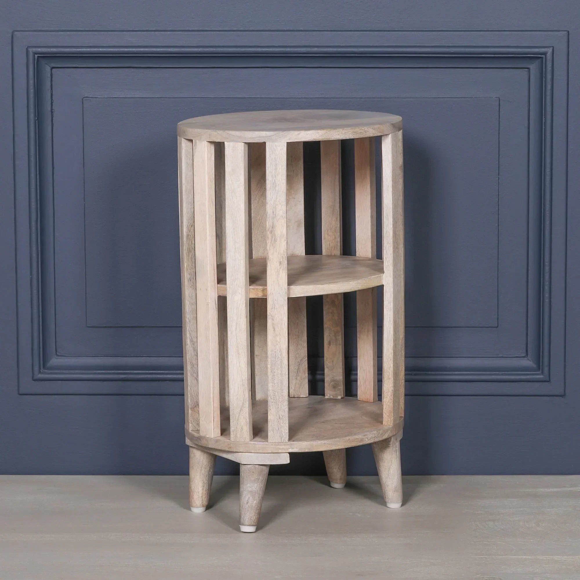 Round wooden side table with vertical slats and two open shelves against blue wall