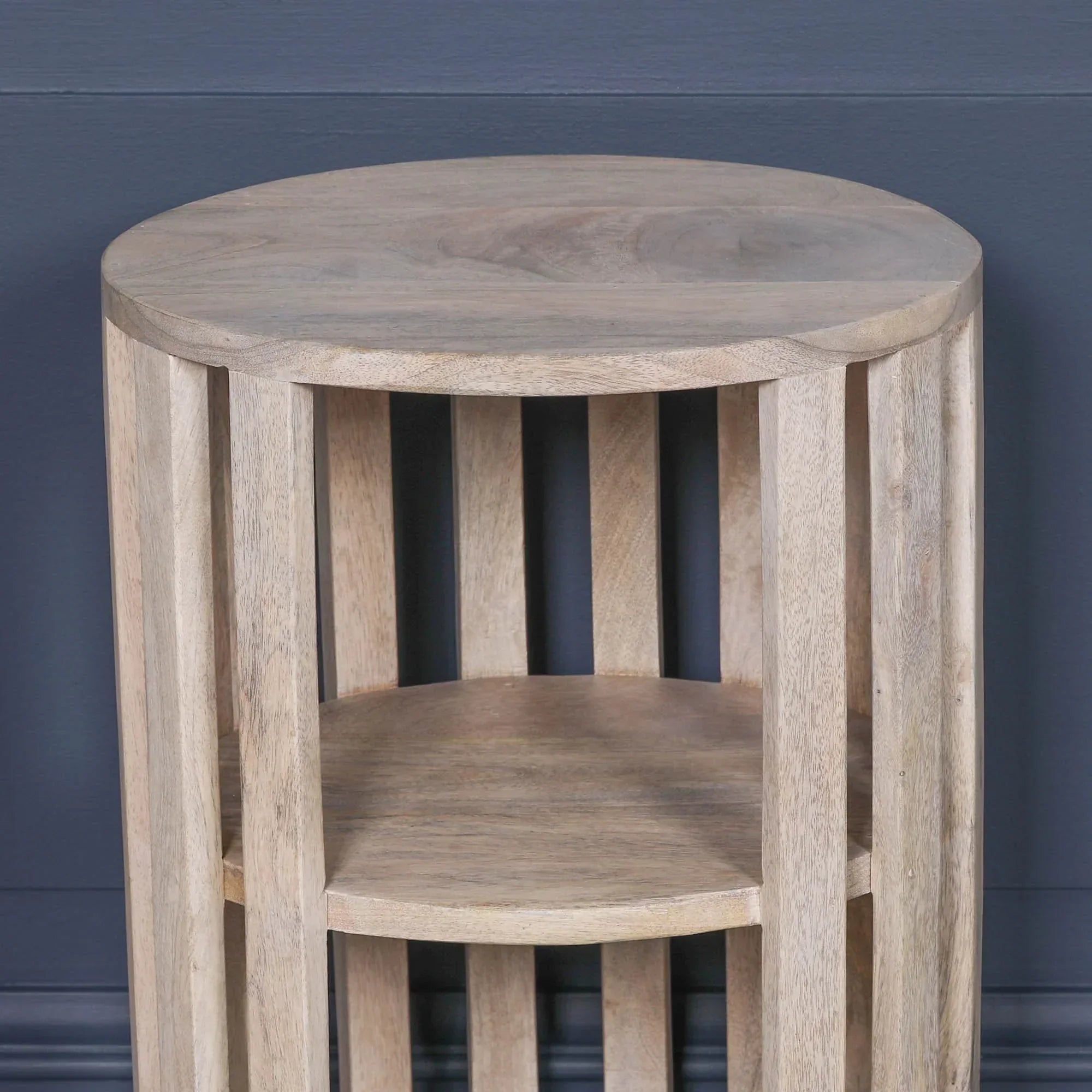 Round wooden side table with slatted frame and lower shelf against dark wall
